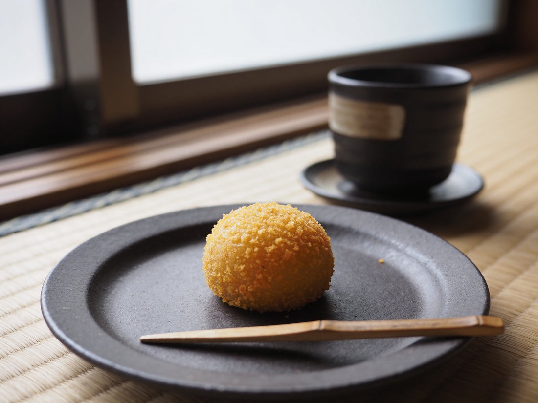 A round Japanese sweet coated in fine crumbs on a dark plate with a wooden pick, a cup of tea behind it