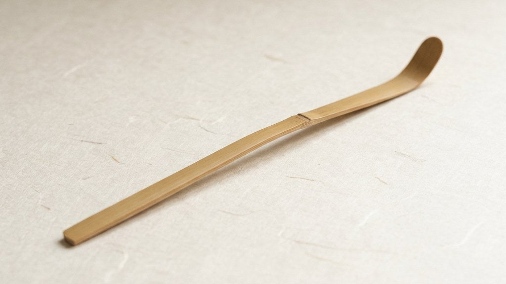A pale bamboo chashaku tea scoop with a single node, lying on white washi paper