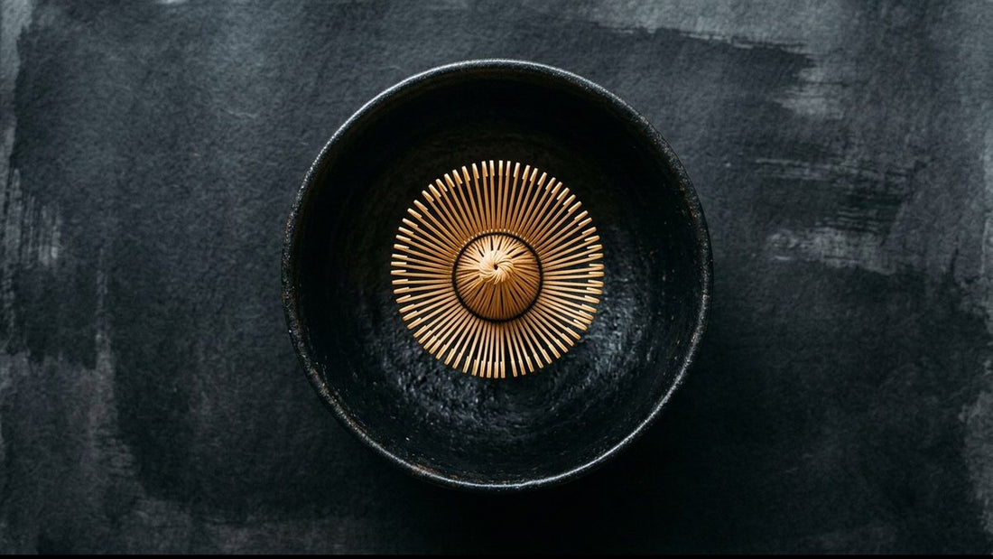 A bamboo matcha whisk resting in the middle of a wide black tea bowl, seen from above, with room on every side