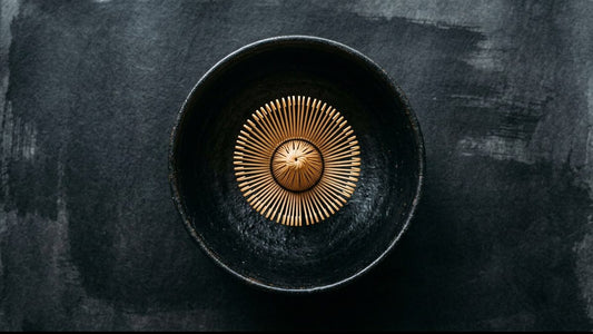 A bamboo matcha whisk resting in the middle of a wide black tea bowl, seen from above, with room on every side