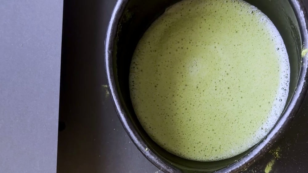Overhead view of a bowl of whisked thin tea in a dark spouted bowl, a few grains of matcha scattered on the dark table around it