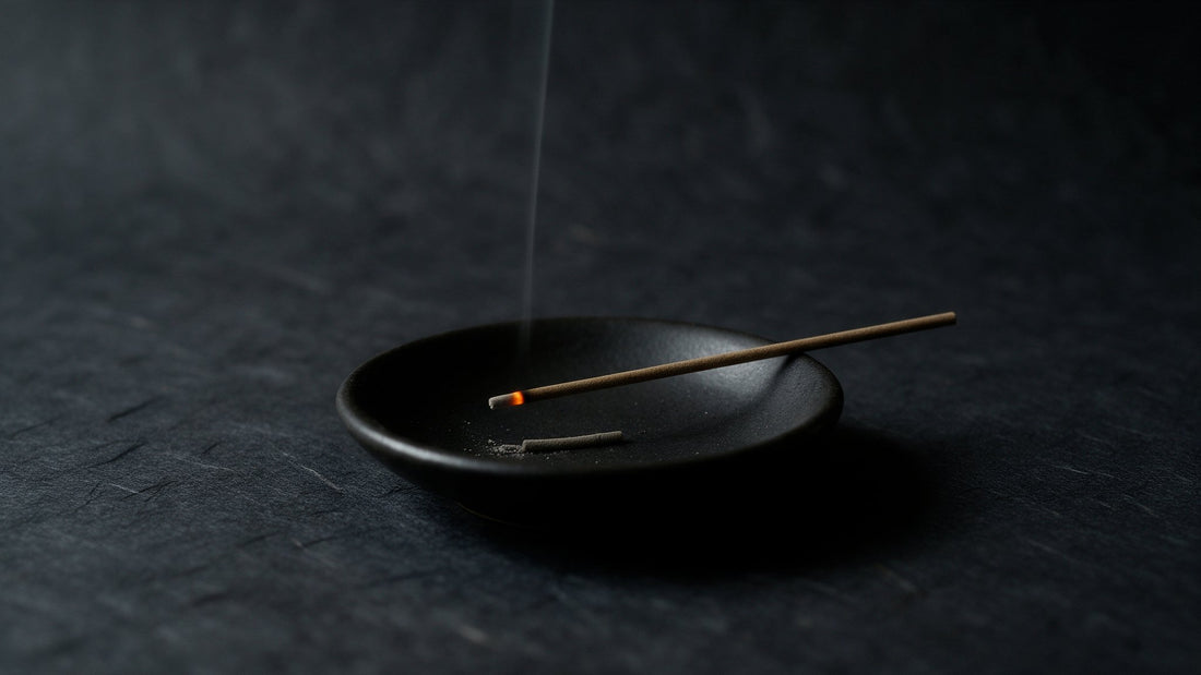 A short Japanese incense stick lying across a small matte black dish, an orange ember at its tip and a single thread of smoke rising, with fallen ash in the dish