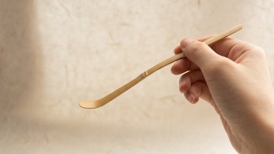 A hand holding a white bamboo chashaku, the node visible partway along the handle, against pale washi paper