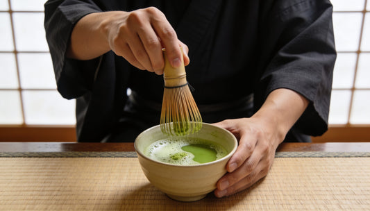 Hands whisking matcha in a bowl, the left hand steadying the bowl, on a tatami mat by a shoji screen