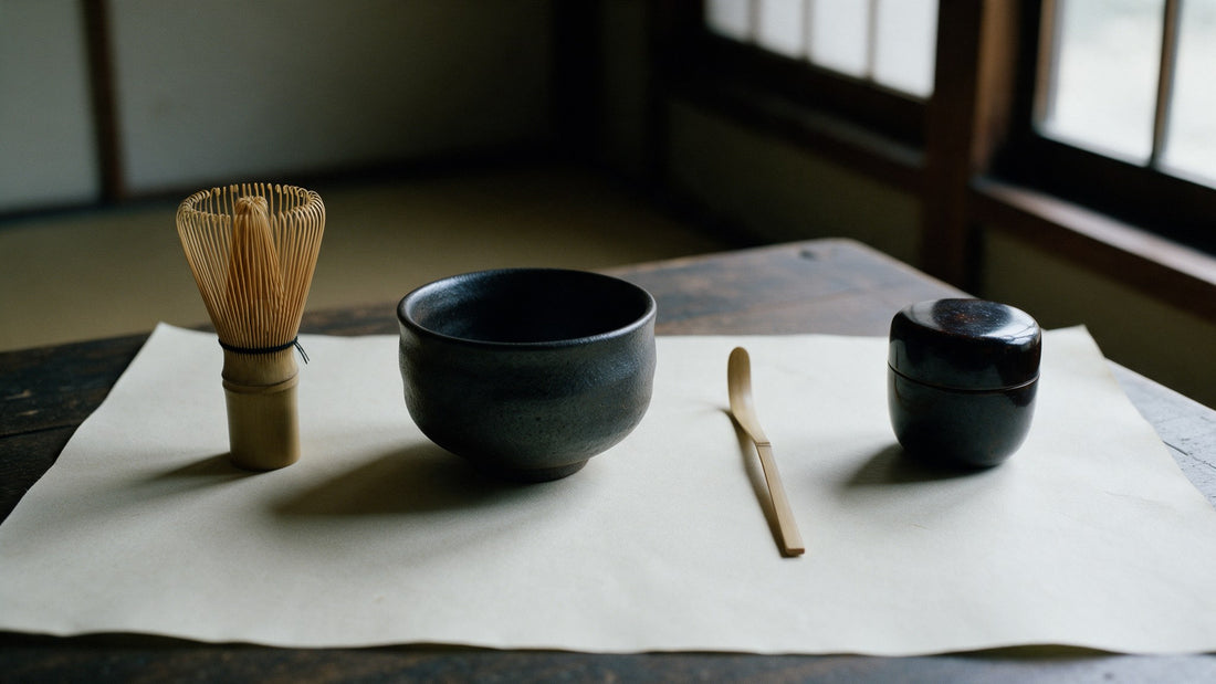 A bamboo matcha whisk, a black Mino-yaki tea bowl, a bamboo tea scoop and a lacquer tea caddy laid out in an even row on washi paper