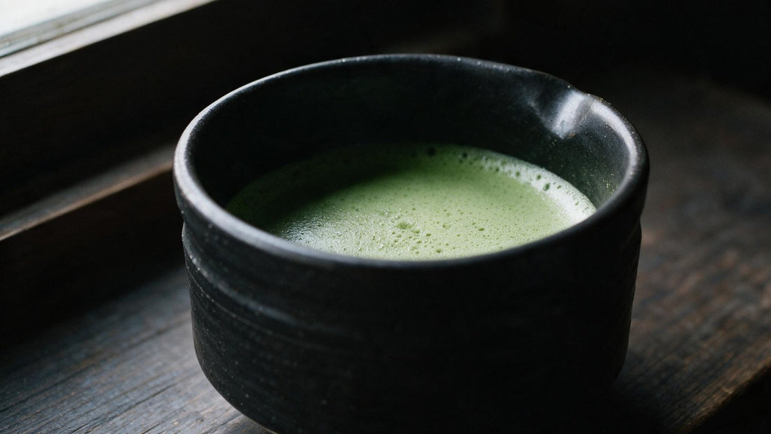 A black tea bowl of whisked matcha on a dark wooden windowsill in soft side light