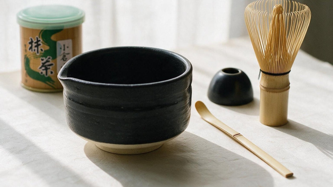 A black tea bowl, bamboo whisk, scoop, whisk holder and a tin of matcha laid out on a pale linen table