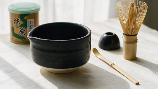 A black tea bowl, bamboo whisk, scoop, whisk holder and a tin of matcha laid out on a pale linen table