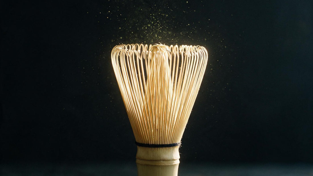 A bamboo tea whisk backlit on a dark surface, its tines catching the light