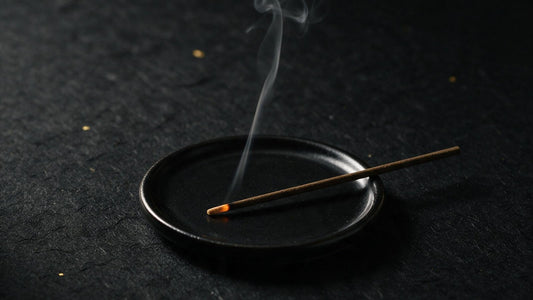A single Japanese incense stick laid across a small black dish, its tip glowing and a thread of smoke rising in a dark room
