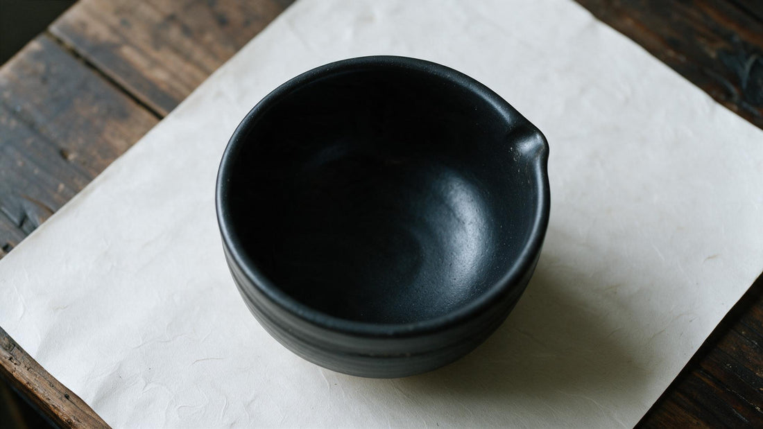 An empty black Mino-yaki matcha bowl on cream washi paper, the rim eased into a low pouring lip at one side