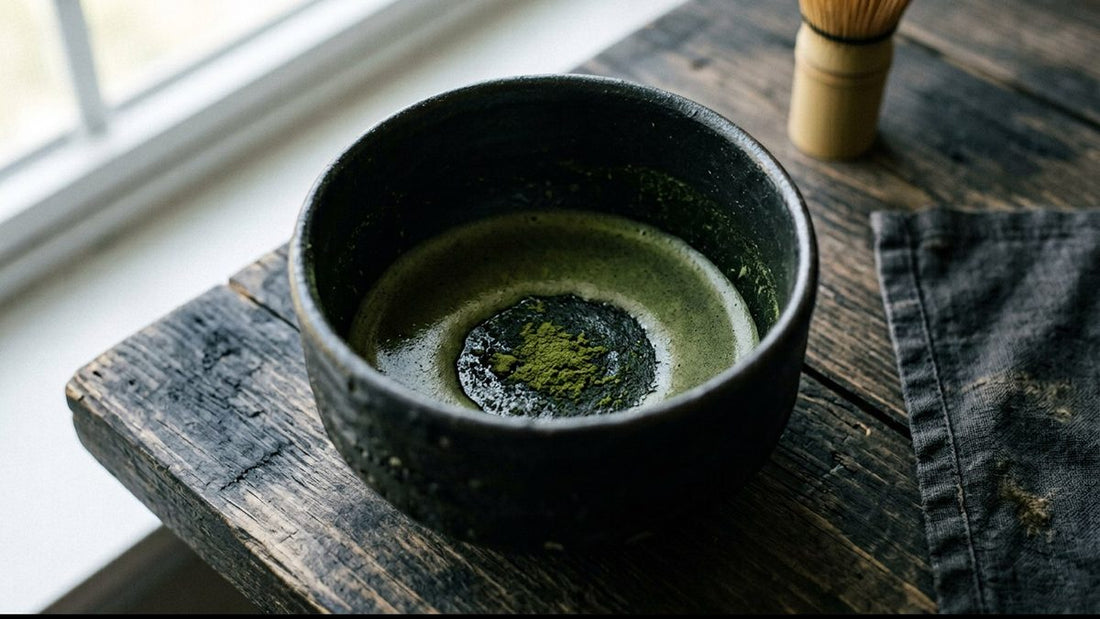 A black tea bowl with a little matcha left in it on a dark wooden table by a window, a bamboo whisk behind
