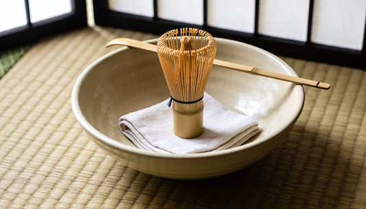 A folded linen chakin in a tea bowl, with a bamboo whisk standing in the bowl and a scoop resting across the rim
