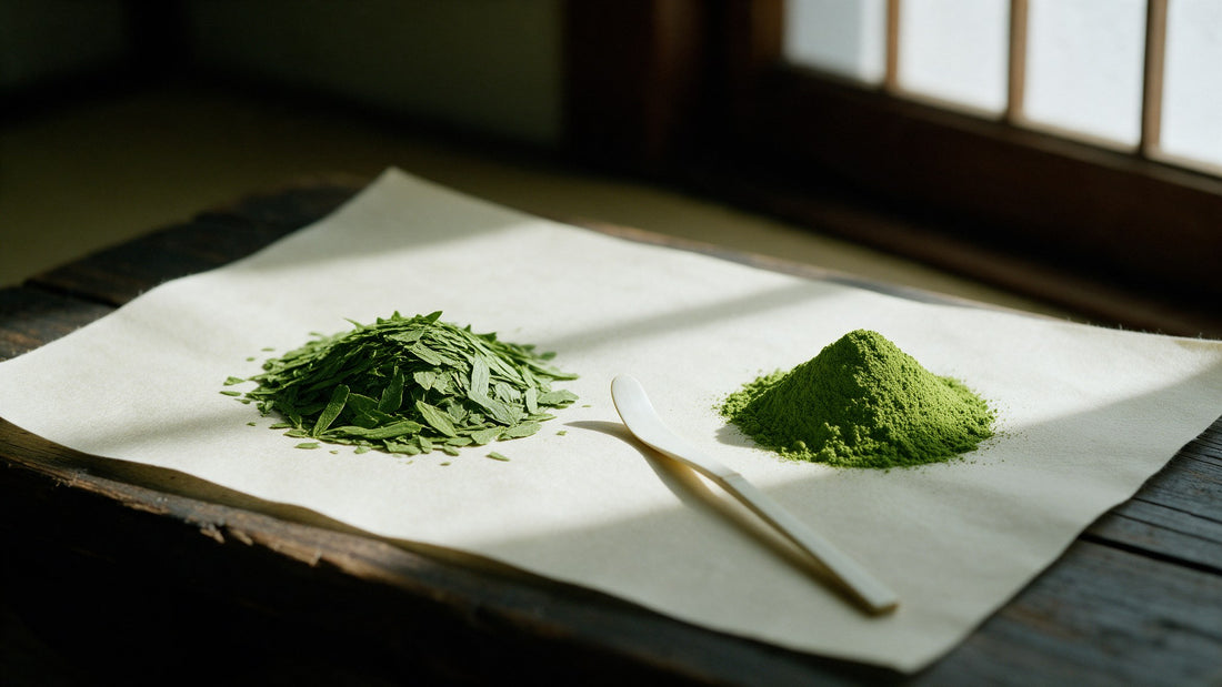 A pile of flat dried tencha leaf flakes beside a mound of fine matcha powder on cream washi paper, a bamboo scoop between them