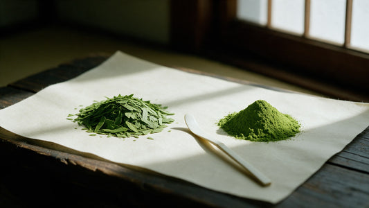 A pile of flat dried tencha leaf flakes beside a mound of fine matcha powder on cream washi paper, a bamboo scoop between them