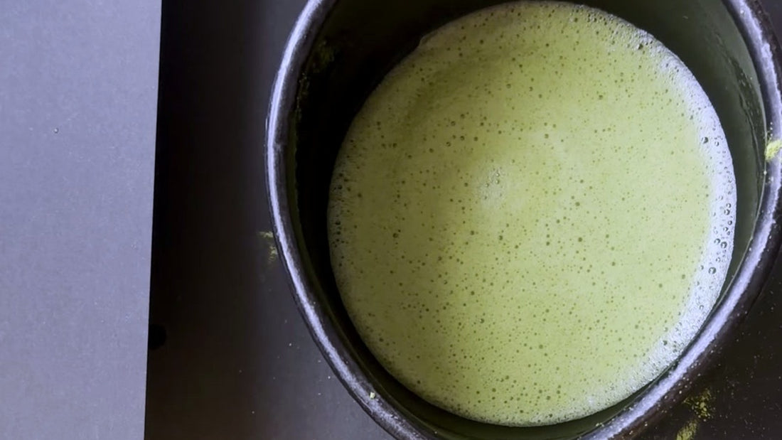 A bowl of whisked matcha seen from above, with loose green powder caught on the rim and the table beside it