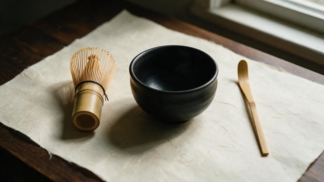 A bamboo chasen, an empty black Mino-yaki tea bowl and a bamboo scoop laid out on cream washi paper beside a window
