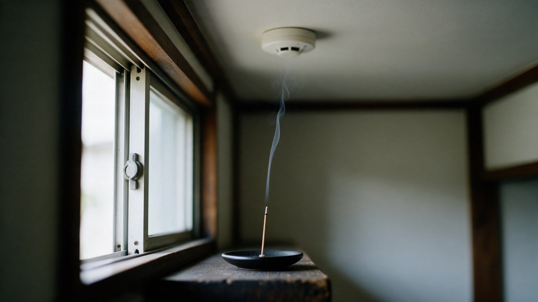 A single Japanese incense stick burning on a black dish by an open window, one thread of smoke rising toward a white smoke alarm on the ceiling