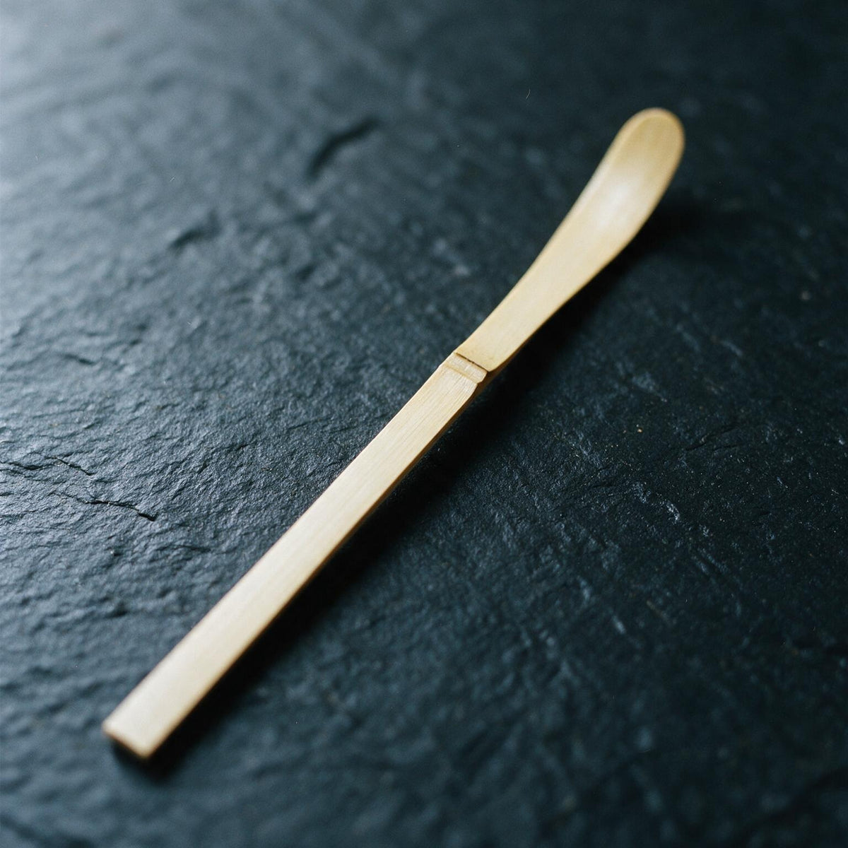 A white-bamboo chashaku on dark slate: a flat strip about 19 cm long and 1 cm wide, with a node and a barely hollowed tip