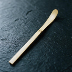 A white-bamboo chashaku on dark slate: a flat strip about 19 cm long and 1 cm wide, with a node and a barely hollowed tip