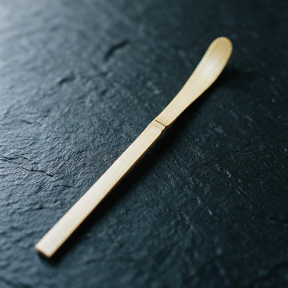 A white-bamboo chashaku on dark slate: a flat strip about 19 cm long and 1 cm wide, with a node and a barely hollowed tip
