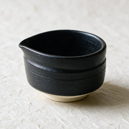 The Mino-yaki chawan: a low, wide, straight-walled bowl in a matte charcoal glaze with a soft pour lip and an unglazed cream foot