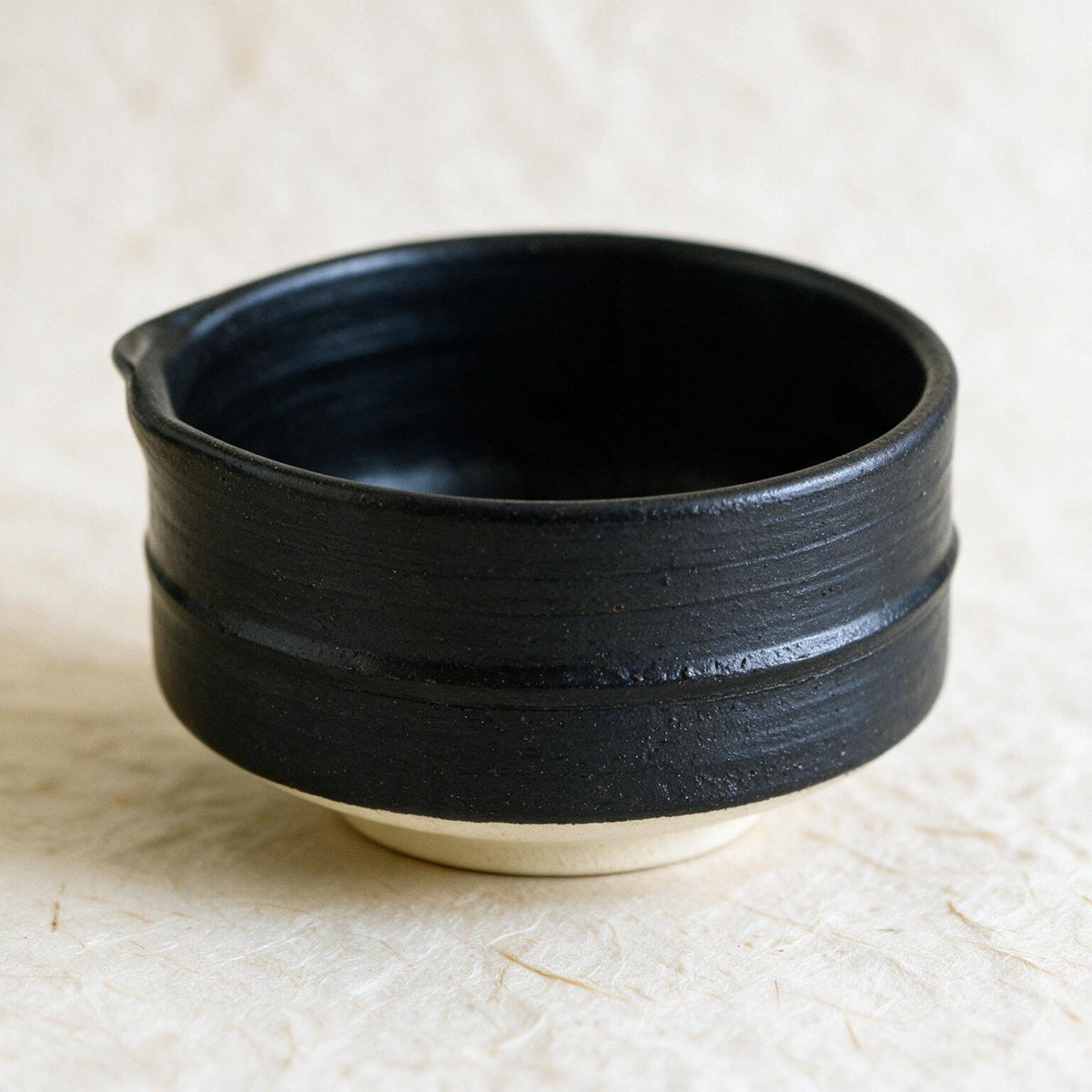 The Mino-yaki chawan: a low, wide, straight-walled bowl in a matte charcoal glaze with a soft pour lip and an unglazed cream foot, seen into the bowl