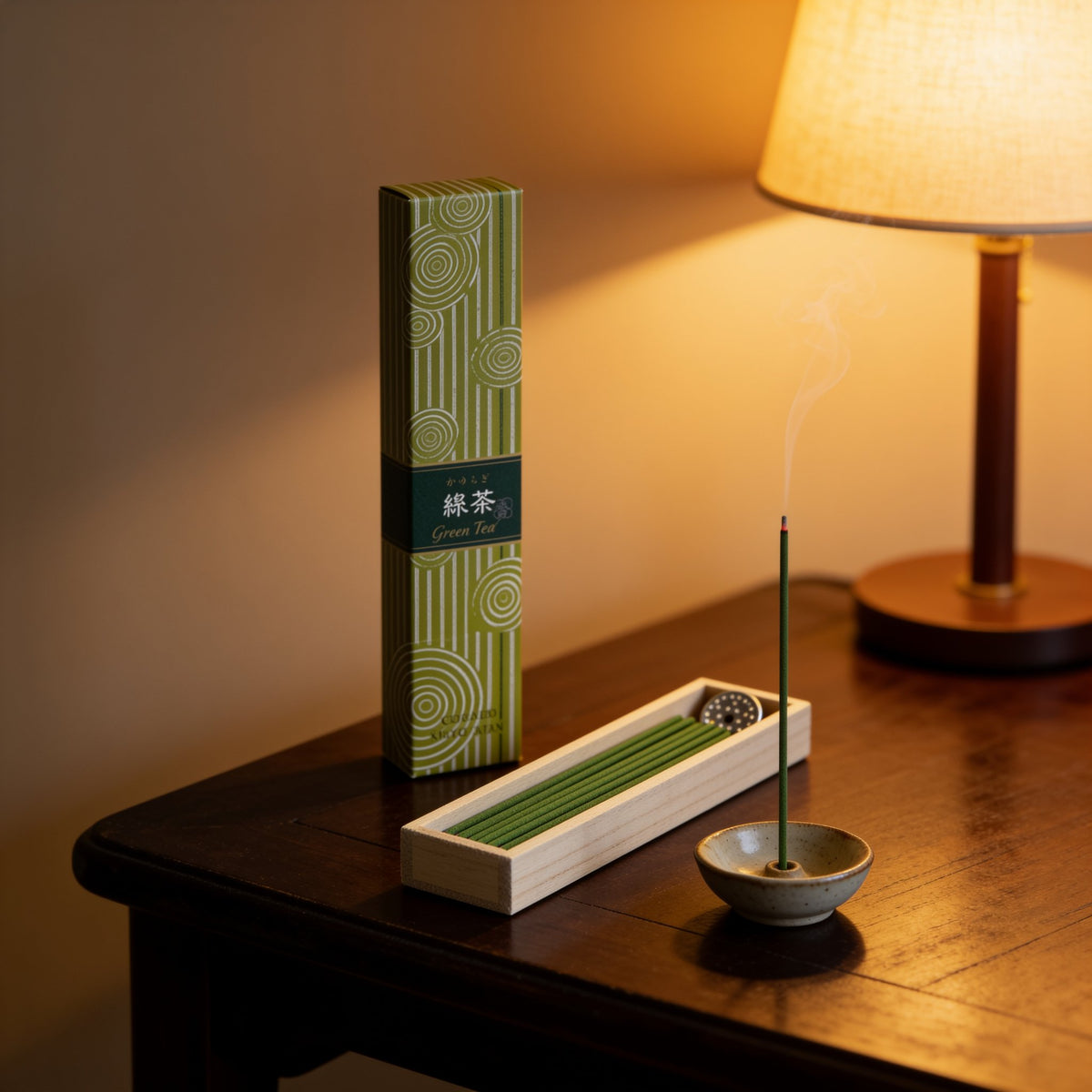 A box of Hyakurakukou Green Tea incense, a tray of green sticks and a lit stick under a table lamp