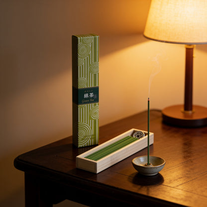 A box of Hyakurakukou Green Tea incense, a tray of green sticks and a lit stick under a table lamp