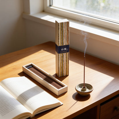 A box of Kayuragi Sandalwood incense, an open tray of sticks and a lit stick beside an open book on a desk by a window