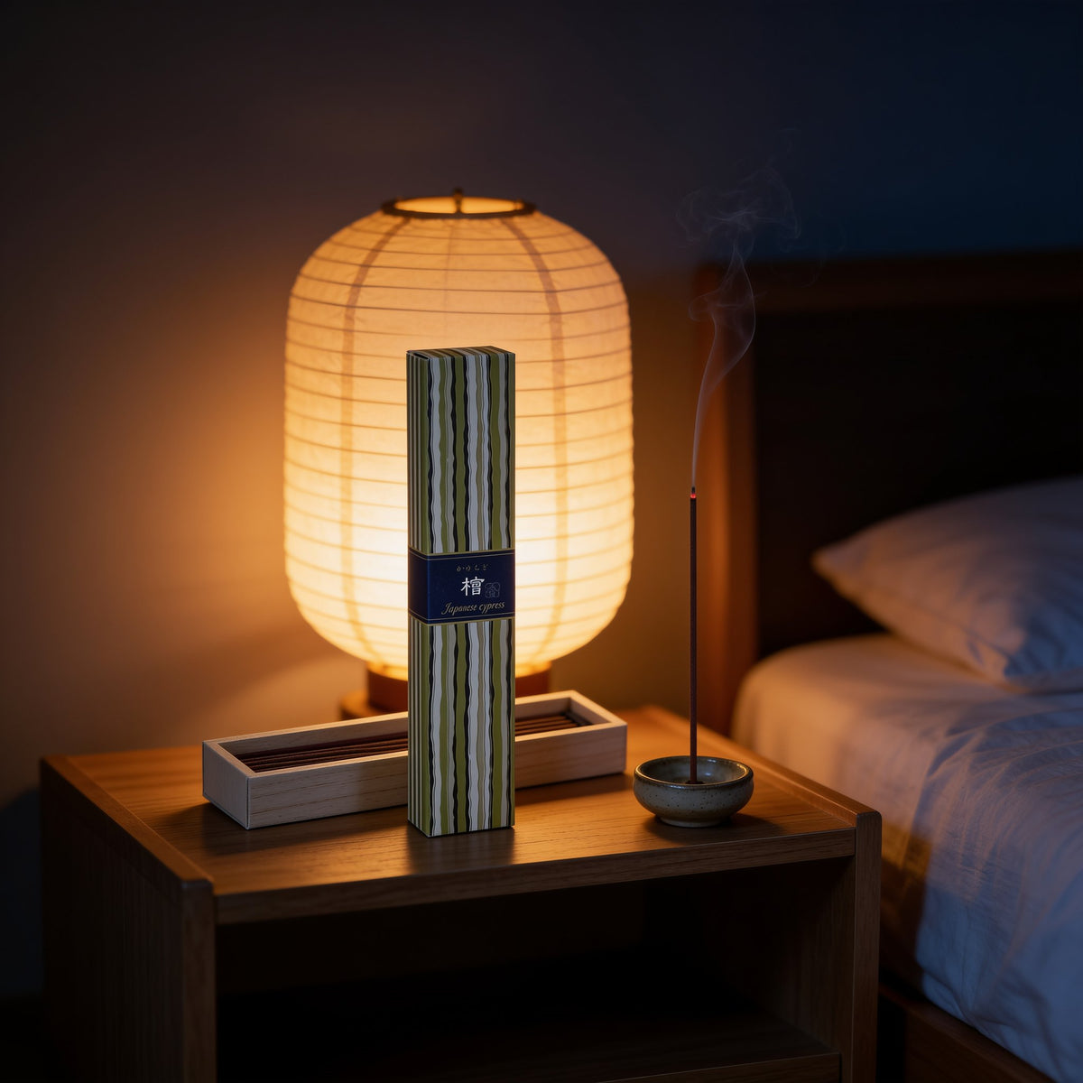 A box of Kayuragi Hinoki incense and a lit stick on a bedside table beside a paper lamp