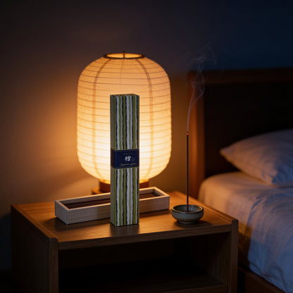 A box of Kayuragi Hinoki incense and a lit stick on a bedside table beside a paper lamp
