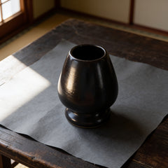 Glossy dark brown-black ceramic kusenaoshi, a matcha whisk shaper, on washi paper. The egg-shaped body is hollow and open at the top, with a narrow waist and a flat round foot