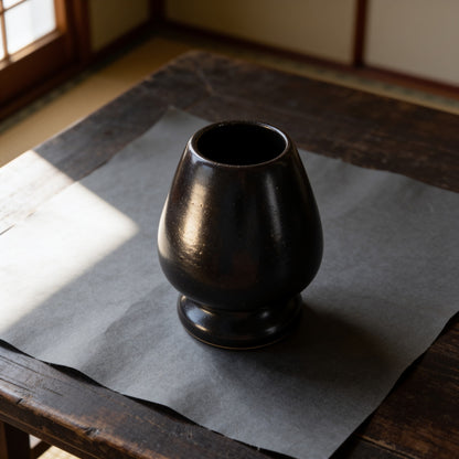 Glossy dark brown-black ceramic kusenaoshi, a matcha whisk shaper, on washi paper. The egg-shaped body is hollow and open at the top, with a narrow waist and a flat round foot