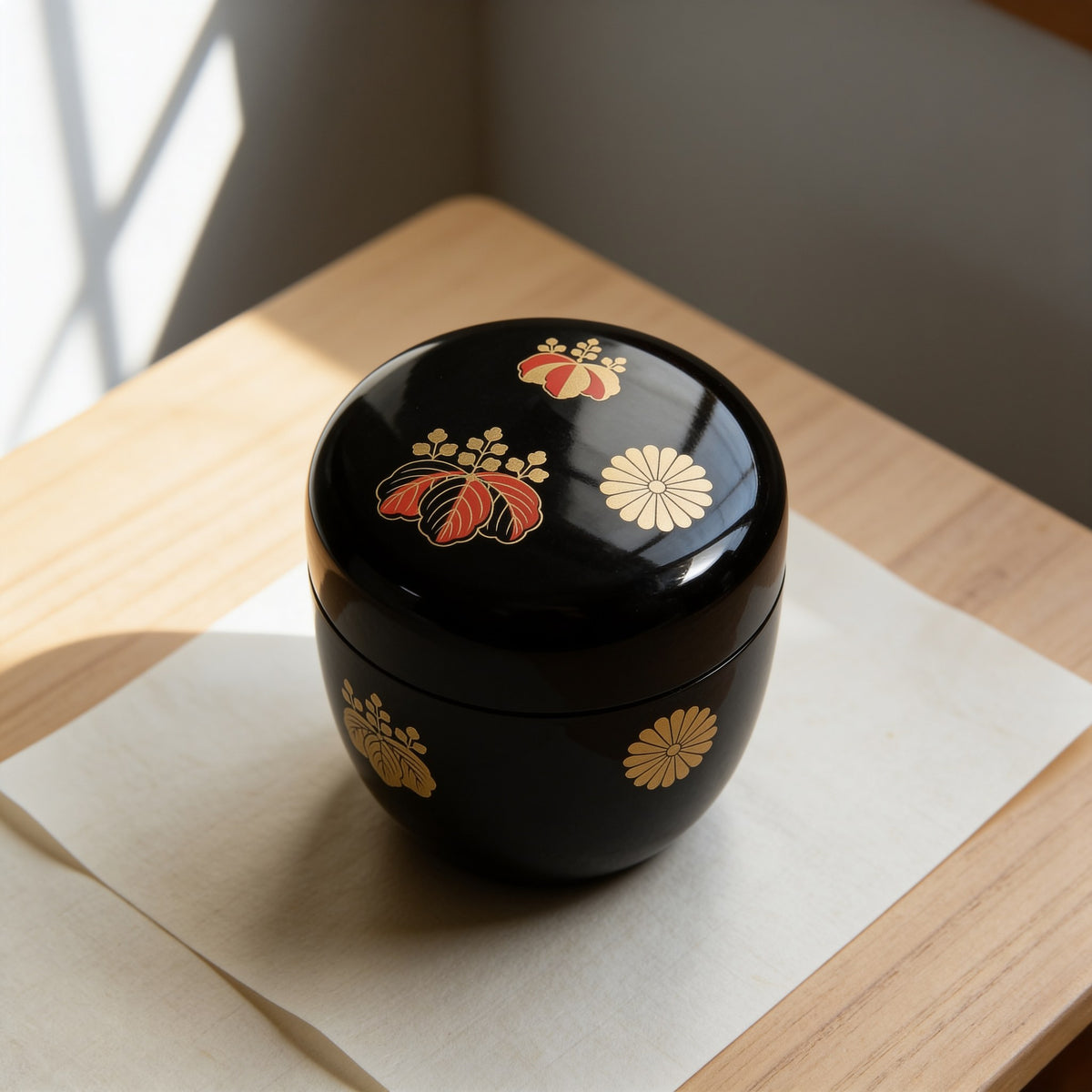 A black lacquered natsume tea caddy with gold and vermilion paulownia crests and a gold chrysanthemum crest, lid closed, on washi paper