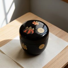 A black lacquered natsume tea caddy with gold and vermilion paulownia crests and a gold chrysanthemum crest, lid closed, on washi paper