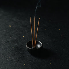 Three incense sticks standing in a small black ceramic holder, one of them lit, a thread of smoke rising